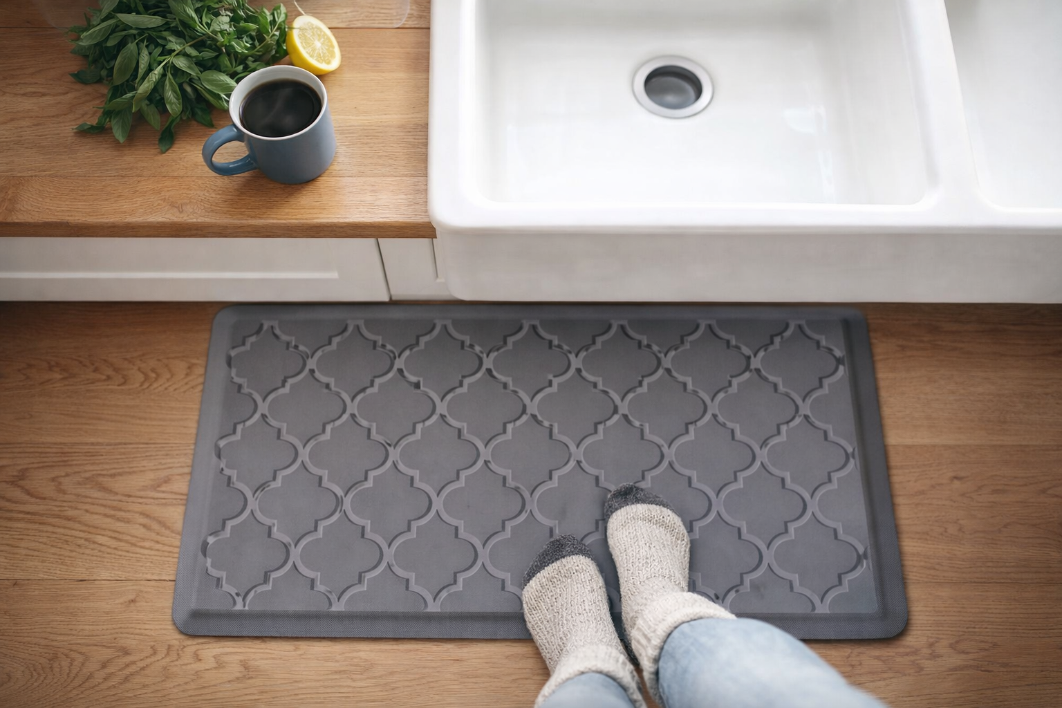 Cozy kitchen scene with a gray anti-fatigue kitchen mat on warm wood flooring in front of a white farmhouse sink, styled with fresh herbs, lemon, coffee, and soft wool socks.